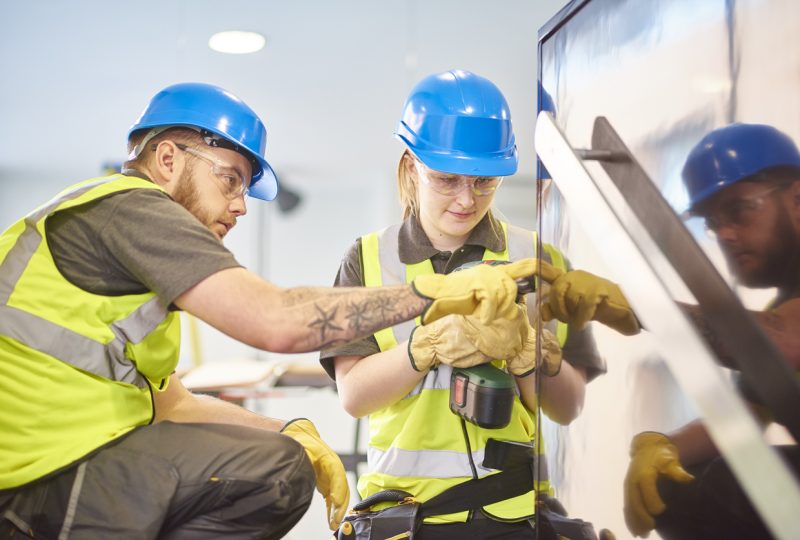 a teenage female apprentice carpenter is installing handrails  in a modern office refurbishment. She is being supervised by her co-worker . She is using a drill to make guide holes for the hand rails  . They are wearing safety workwear.